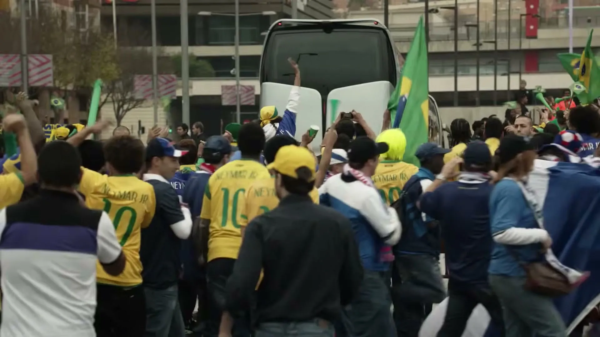 A crowd of excited soccer fans, many wearing yellow and green jerseys, cheer and wave flags around a white bus on a city street.