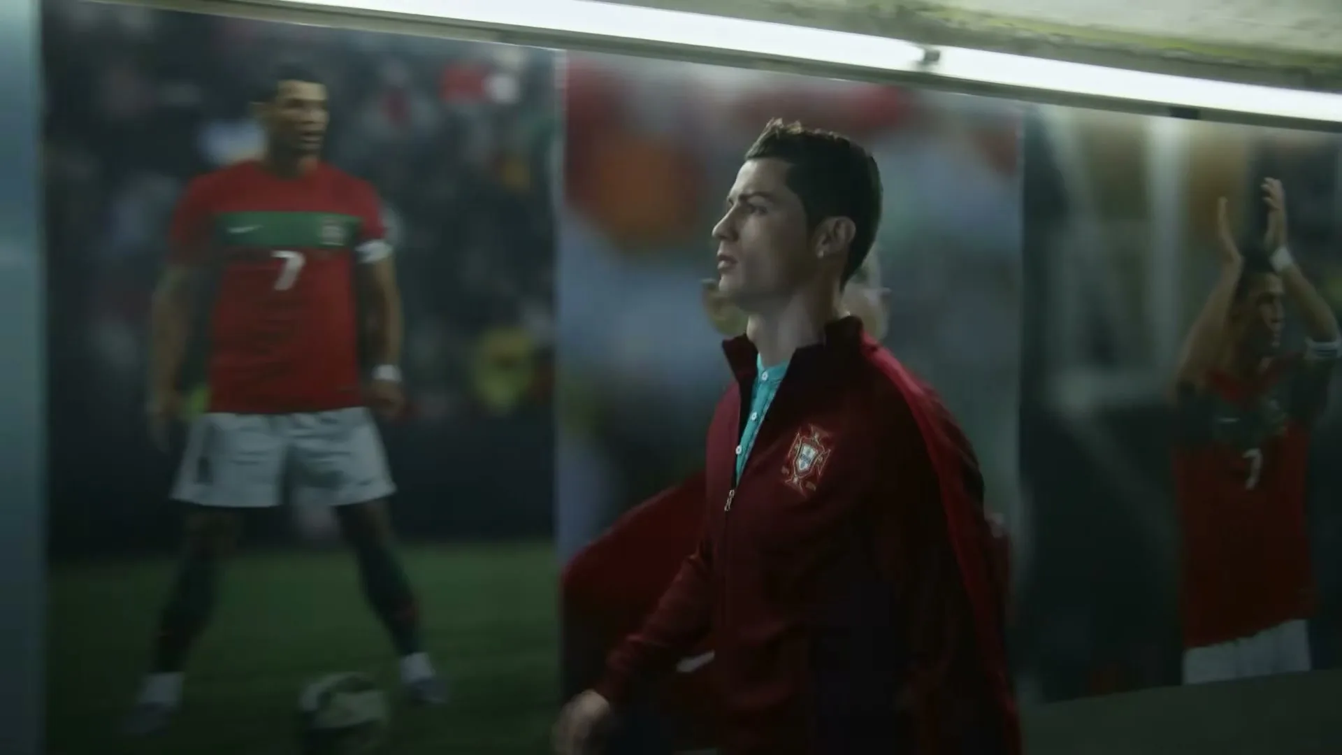 A male soccer player in a red tracksuit walks past large posters of himself in a stadium tunnel.
