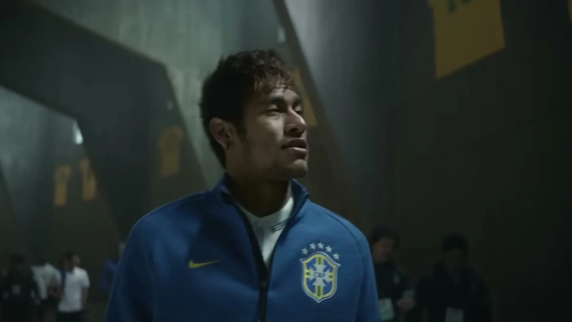 Neymar Jr., a male soccer player in his 20s, wears a blue and yellow Brazil national team jacket, looking up and around in a stadium tunnel.