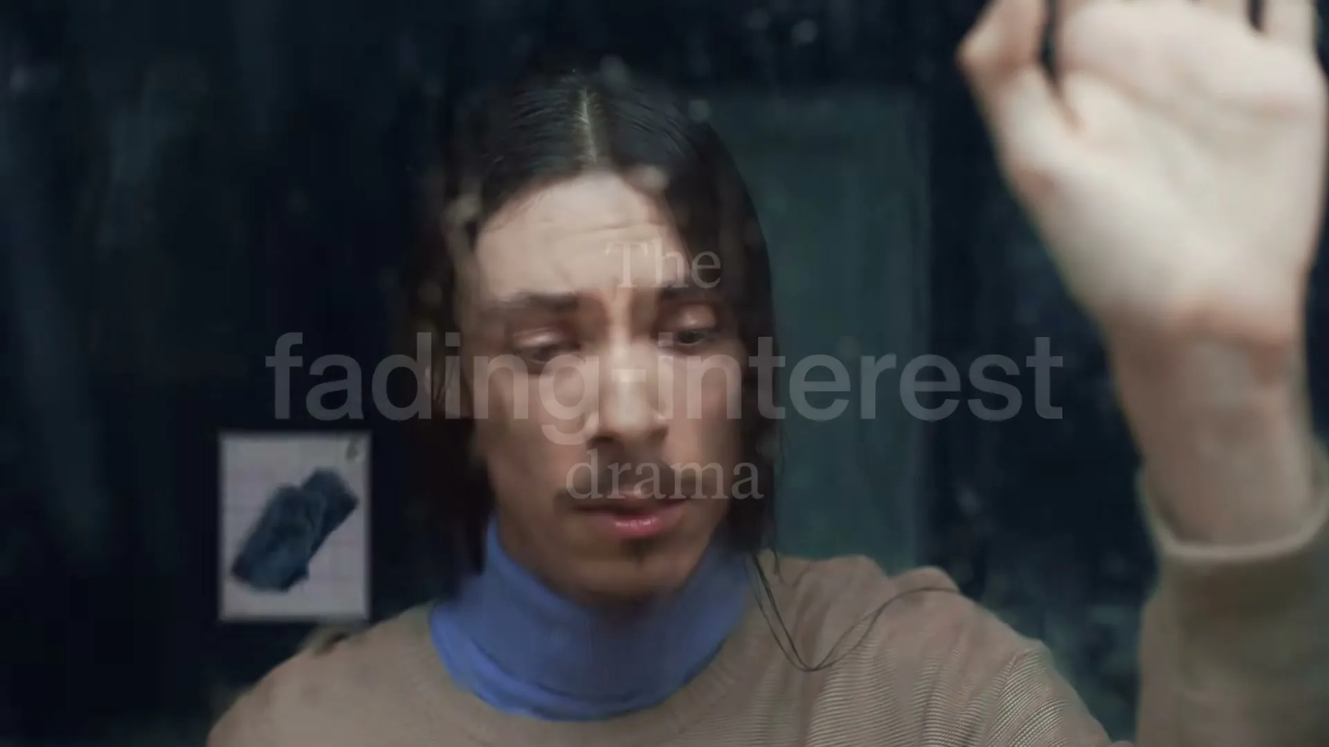 A man with long hair and a mustache looks at a phone screen with a pair of jeans and a message, his hand pressed against a rainy window.