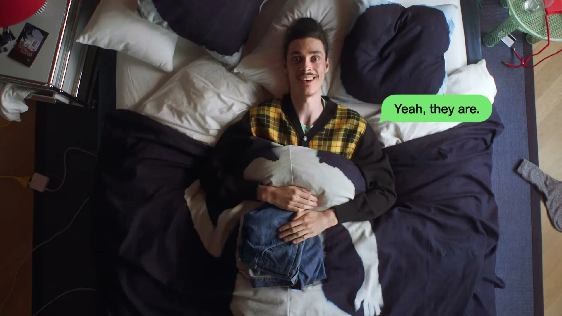 A young man with a mustache lies in bed, holding a pillow and a pair of jeans, then opens his eyes and smiles, with a text bubble appearing.