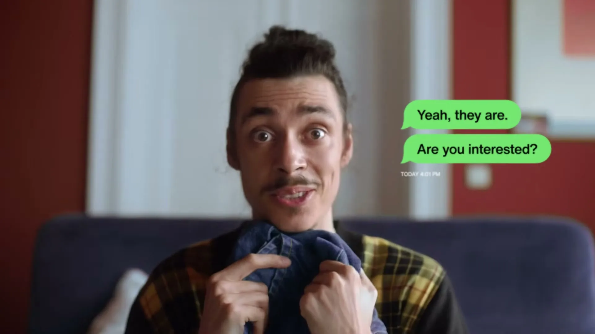 A man with a mustache and bun sits on a couch, holding up a piece of denim clothing, with text messages floating next to him.