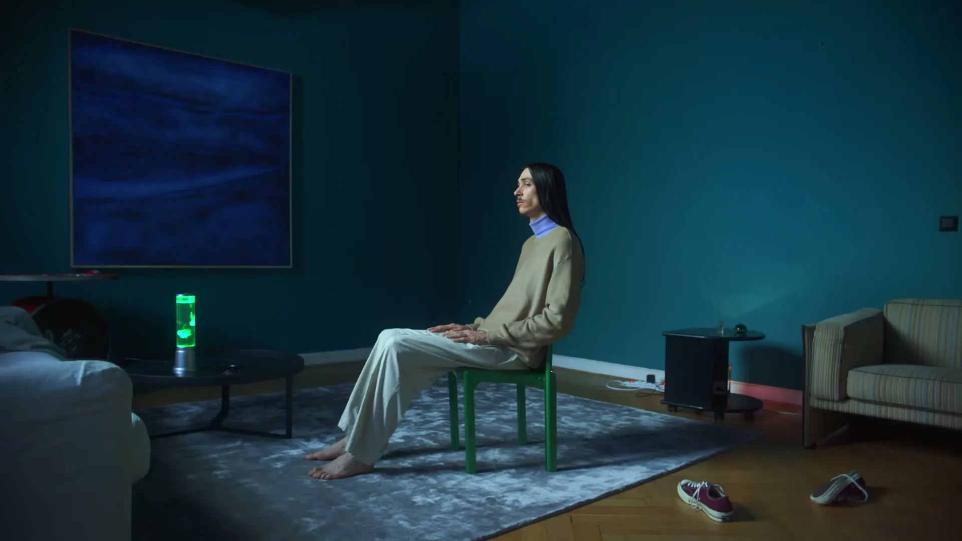 A person with long dark hair sits barefoot on a green chair in a teal room, turning their head to look at the camera.