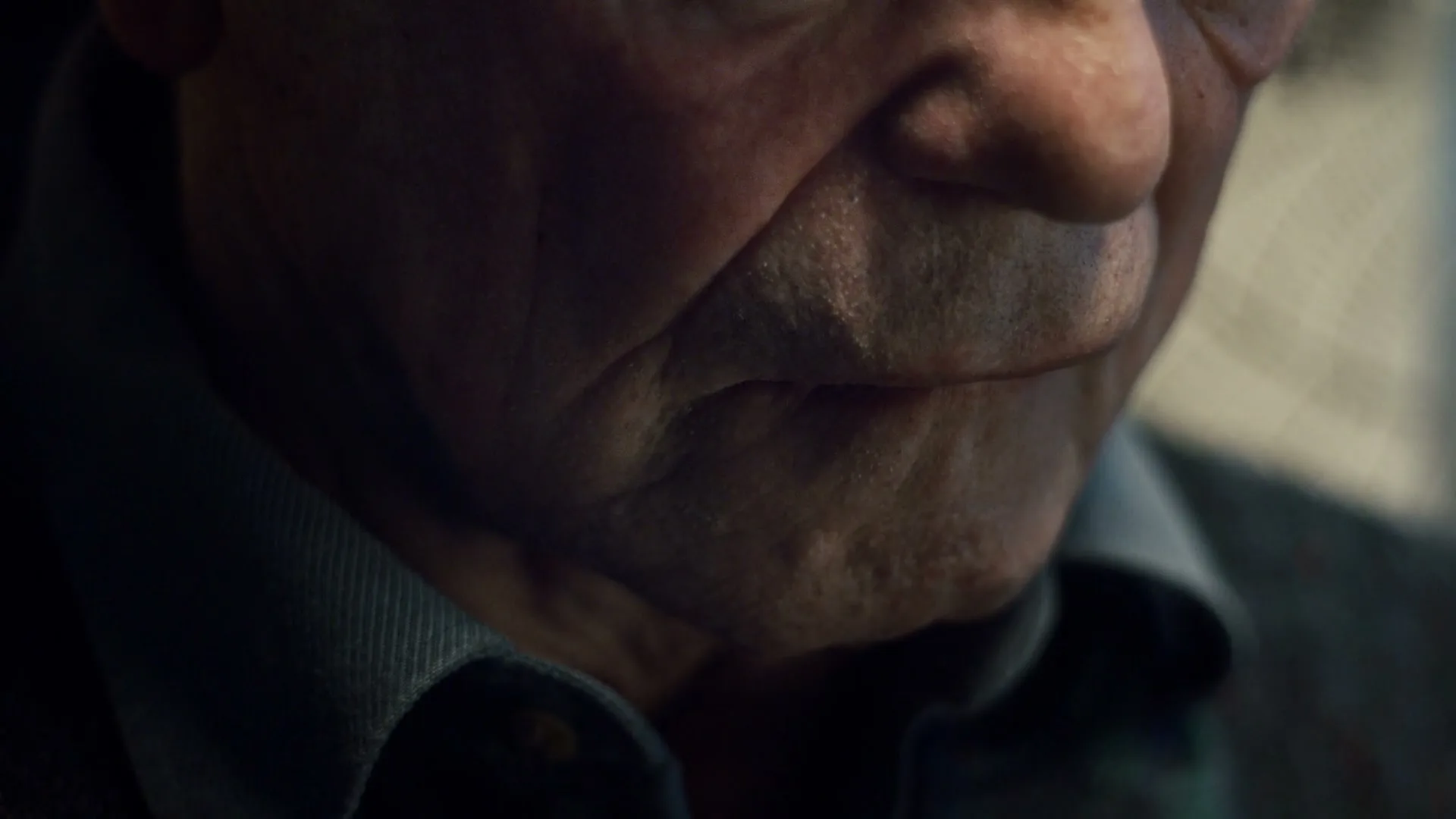 A close-up shot of an older man's mouth and chin, showing his wrinkled skin, as he looks down in a dimly lit interior.