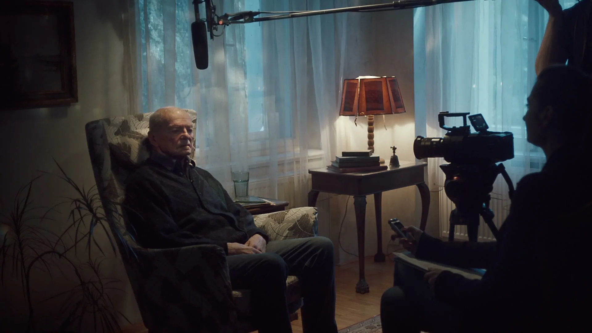 An elderly man sits in an armchair, being interviewed by a person holding a camera in a dimly lit room with a window.