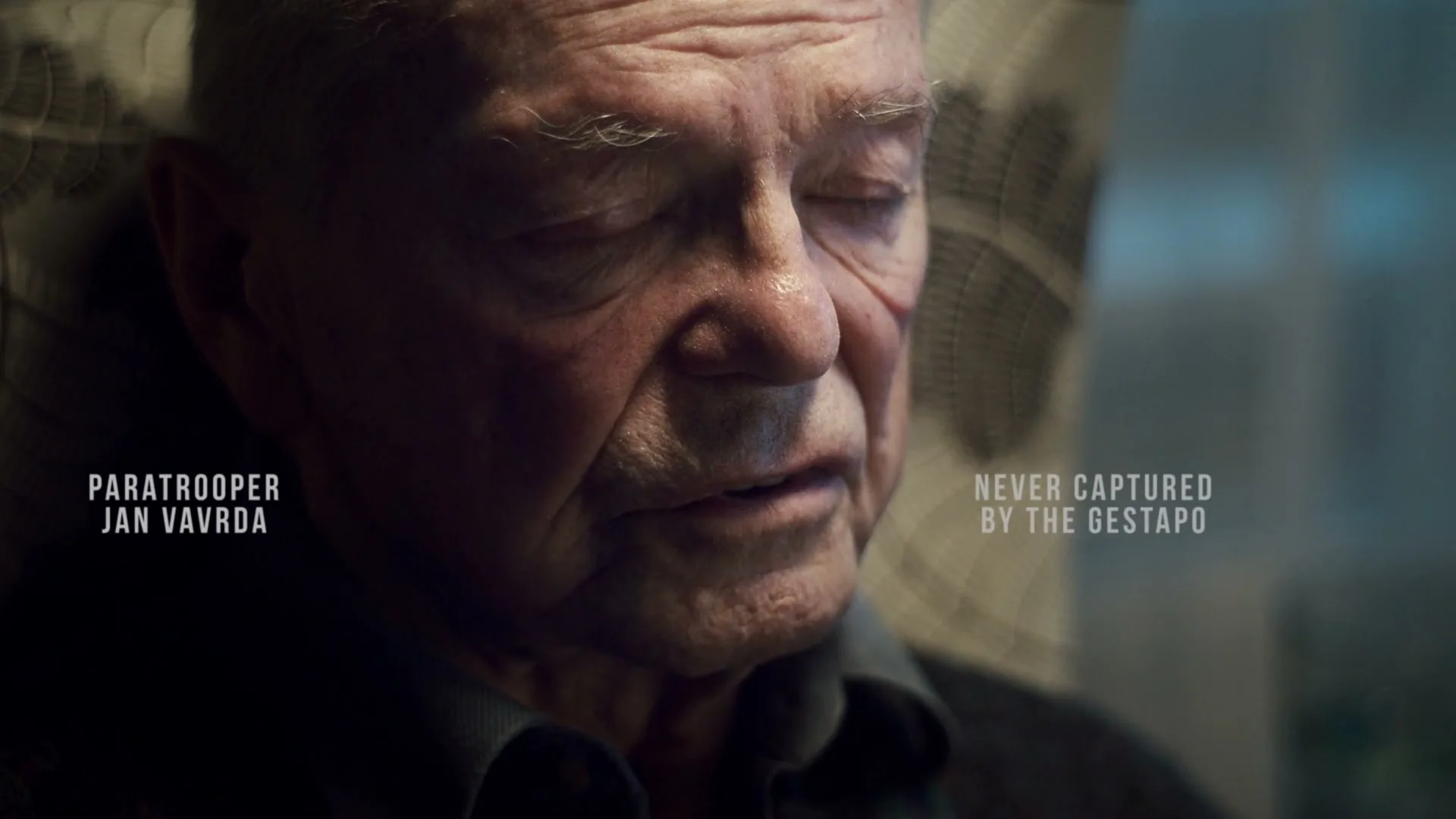 An elderly man with white hair and a blue shirt sits in a chair, looking down with a somber expression in an interior setting.