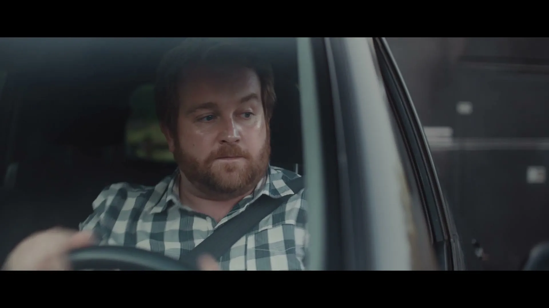 A man with a beard and plaid shirt drives a car, looking intently to his right.
