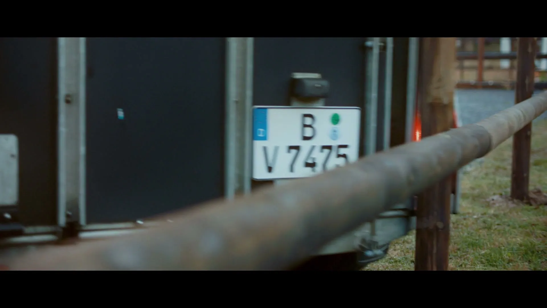 A close-up shot of a German license plate "B V 7475" on the back of a moving vehicle, with a blurred wooden fence in the foreground.