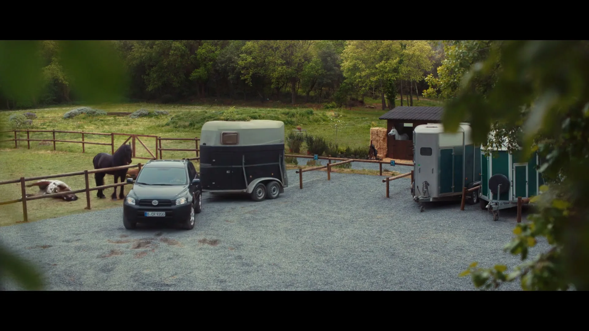 A black SUV with a horse trailer attached is parked on a gravel driveway next to a fenced pasture with horses and a small stable.