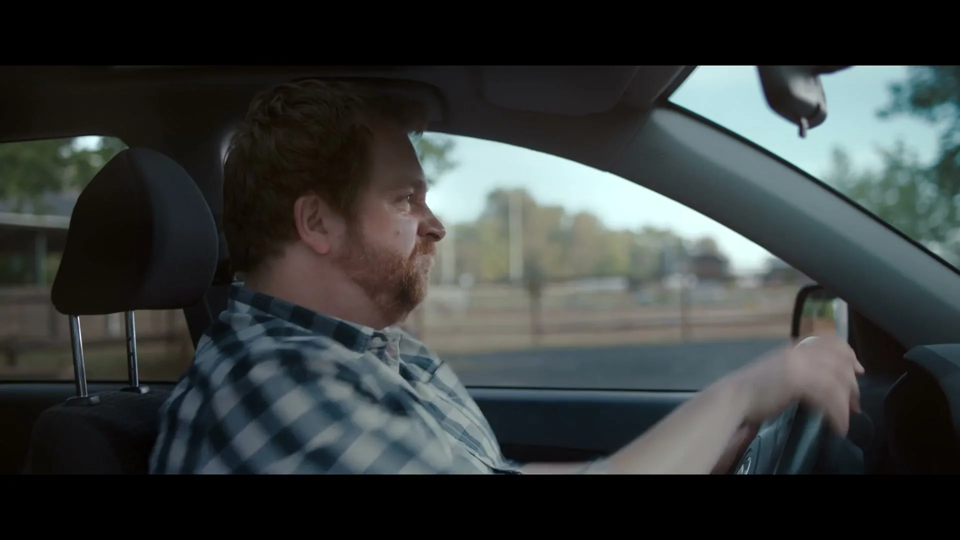 A man with a beard, wearing a plaid shirt, drives a car, looking straight ahead with a neutral expression.