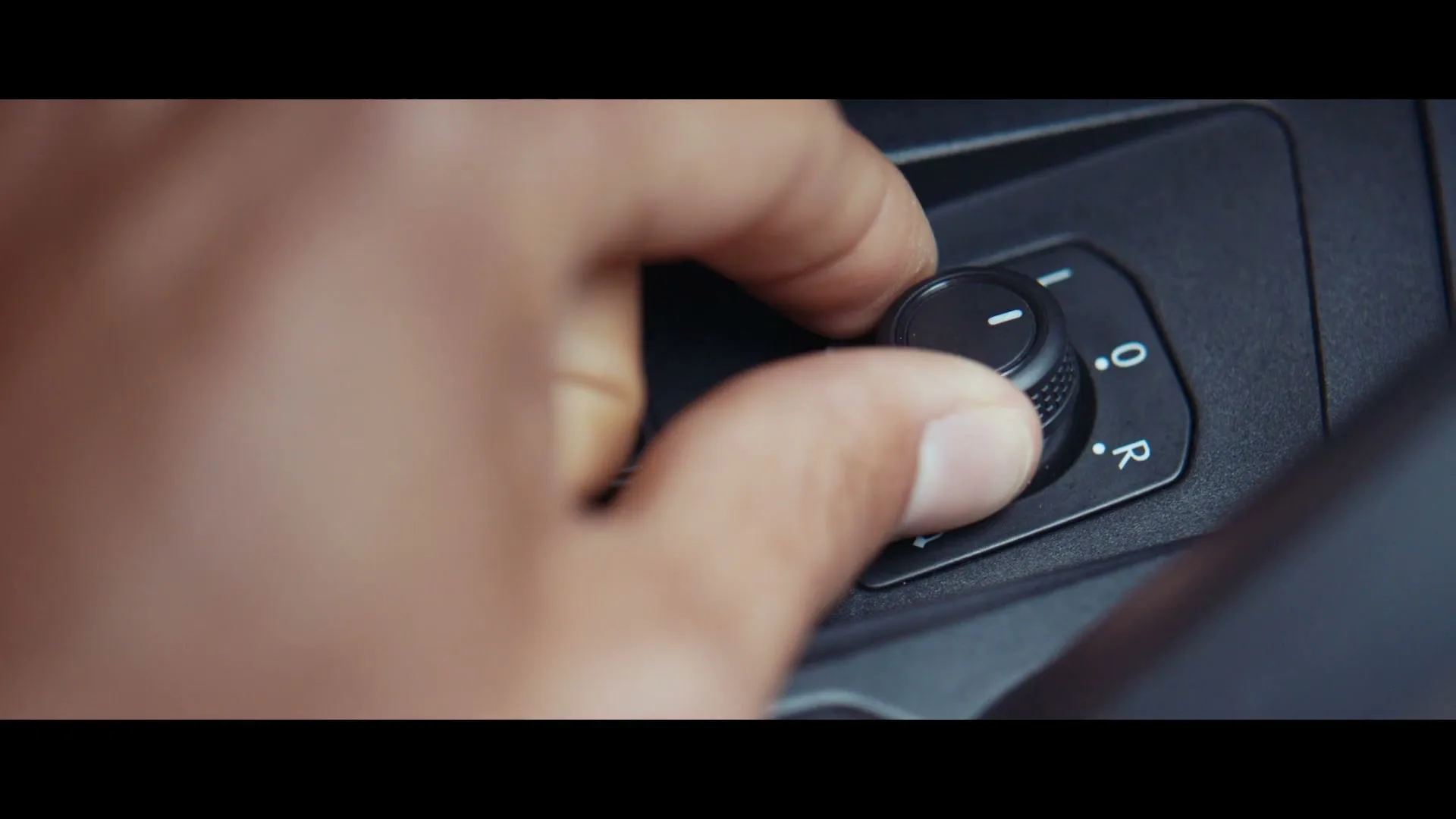 A person's hand adjusts a black knob on a car's interior panel, with letters "L" and "R" visible, in a close-up shot.