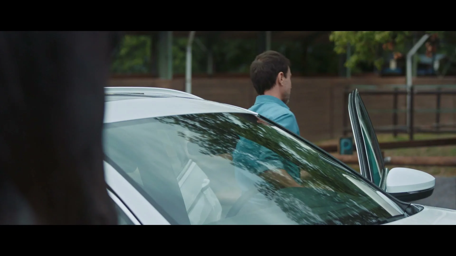 A man in a blue shirt gets out of a white car, looking back at someone off-screen in an outdoor setting.