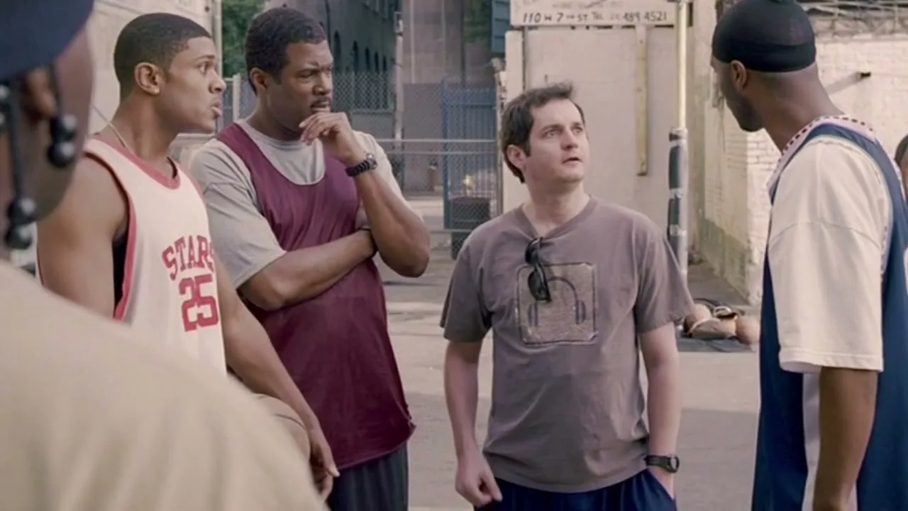 Four men, three Black and one white, stand on an outdoor basketball court, talking and gesturing, with buildings in the background.