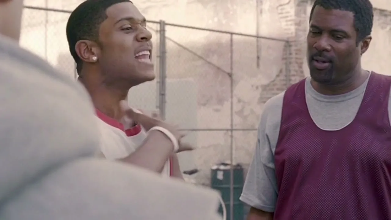 Two men, one in a white and red basketball jersey and another in a grey t-shirt and maroon basketball jersey, are talking on an outdoor basketball court.