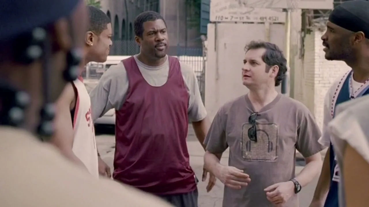 A group of five men, some in basketball jerseys, stand in a circle on a city street, looking at and talking to each other.