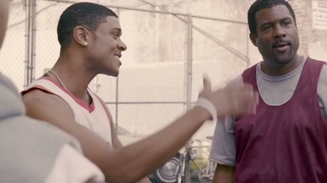 Two men in basketball jerseys talk on an outdoor court, one gesturing with his hand, during the day.