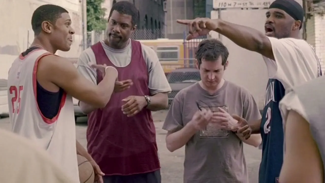 Four men on a basketball court, three of them in basketball jerseys, are discussing something, with one man pointing and another holding a basketball.