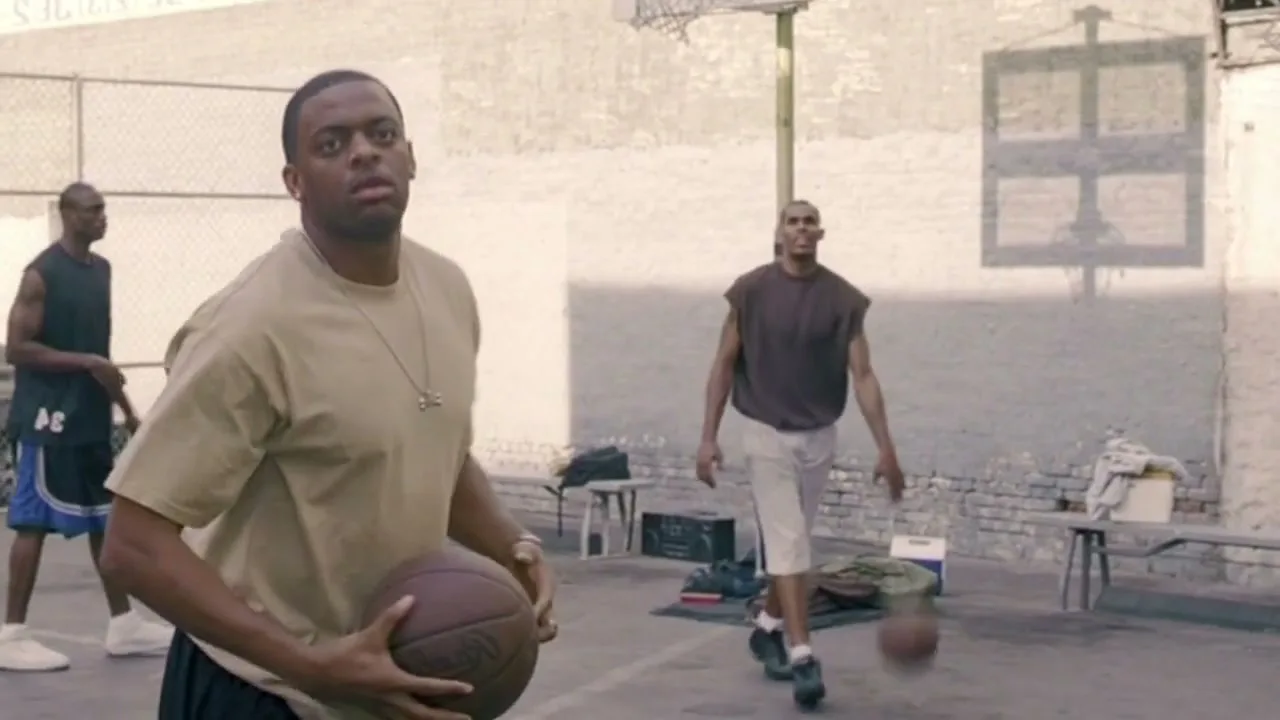 A man in a tan t-shirt holds a basketball and looks over his shoulder at other men playing basketball on an outdoor court.