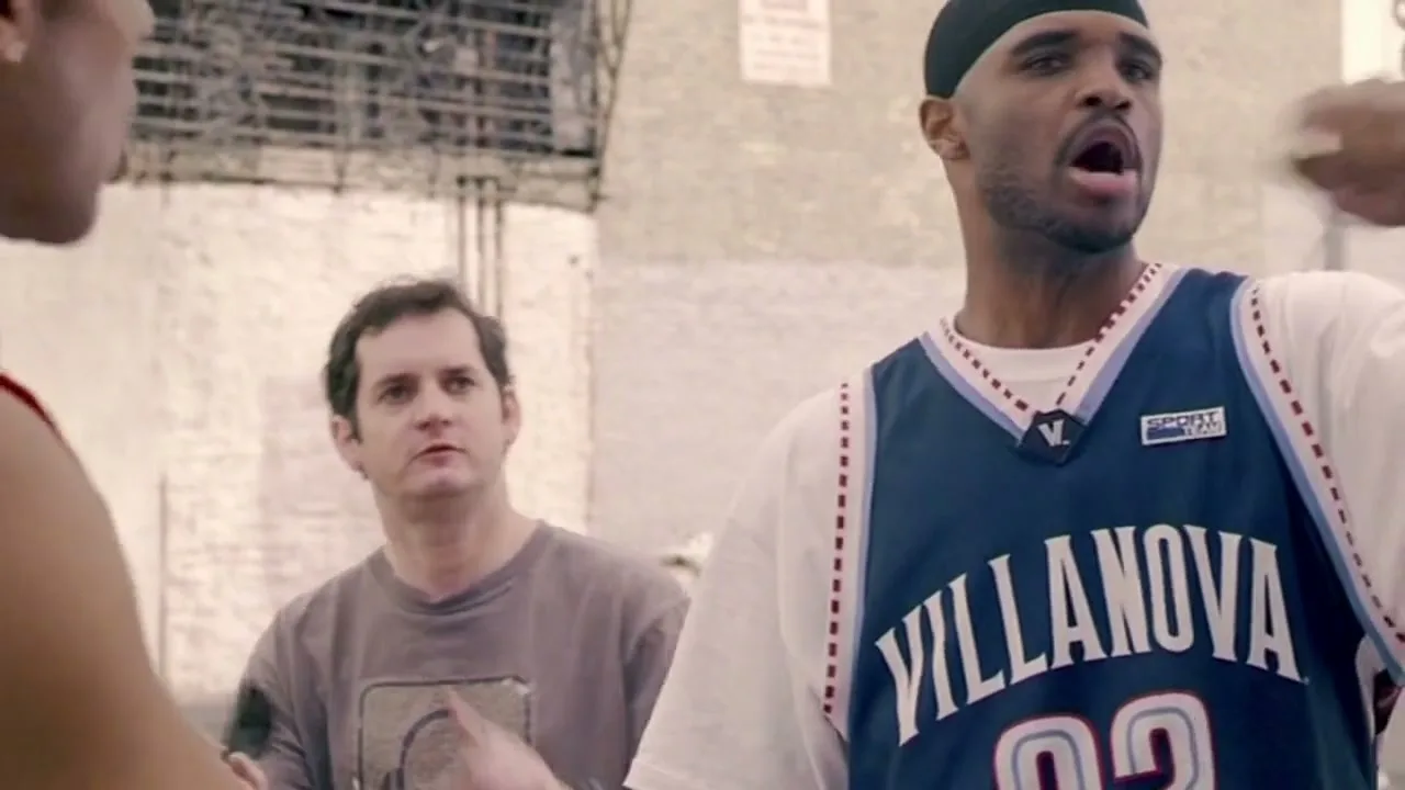 A Black man in a basketball jersey argues with an unseen person, while a white man in a t-shirt stands behind him on an outdoor basketball court.