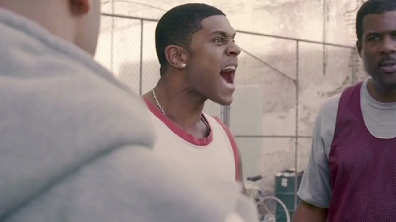 A young Black man in a white and red basketball jersey yells at an unseen person, while an older Black man stands next to him on an outdoor basketball court.