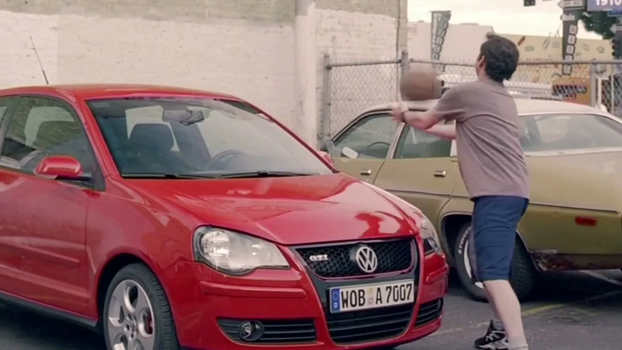 A man in a t-shirt and shorts dribbles a basketball next to a red Volkswagen car in an outdoor parking lot.