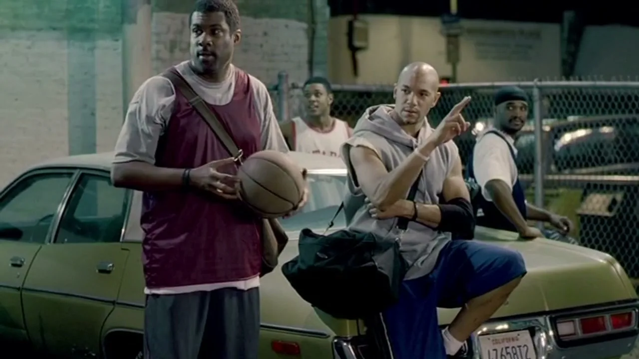 Four men in basketball attire stand and sit around a green car at night, looking and pointing off-screen.