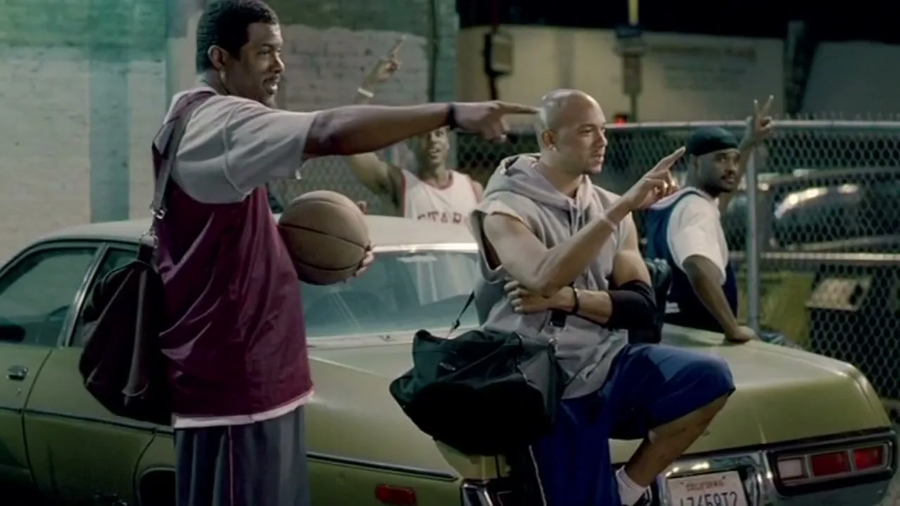 A group of men, dressed in casual sportswear, stand around a parked car, pointing and reacting to something off-screen at night.