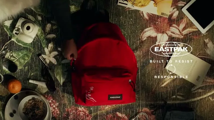 A person's hands are seen from an overhead perspective, packing a red Eastpak backpack on a floral rug surrounded by various personal items.