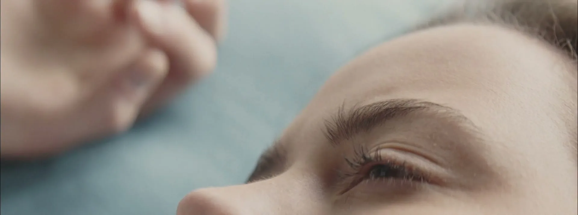 A young woman's eye slowly opens, revealing her pupil, as she lies on a light blue surface.