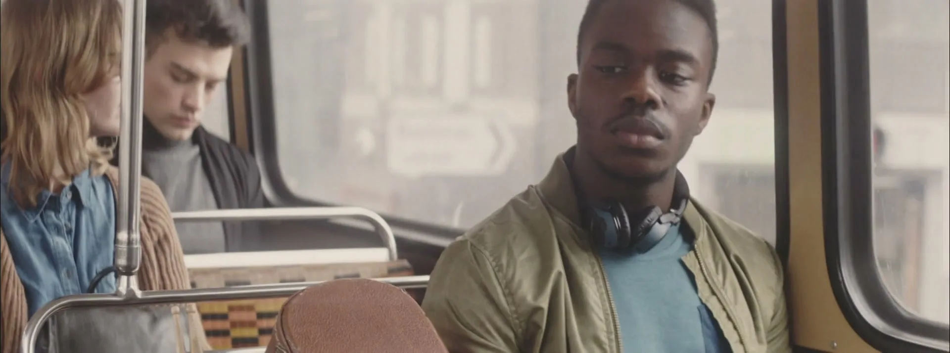 A young Black man with headphones around his neck sits on a bus, looking out the window with a neutral expression.