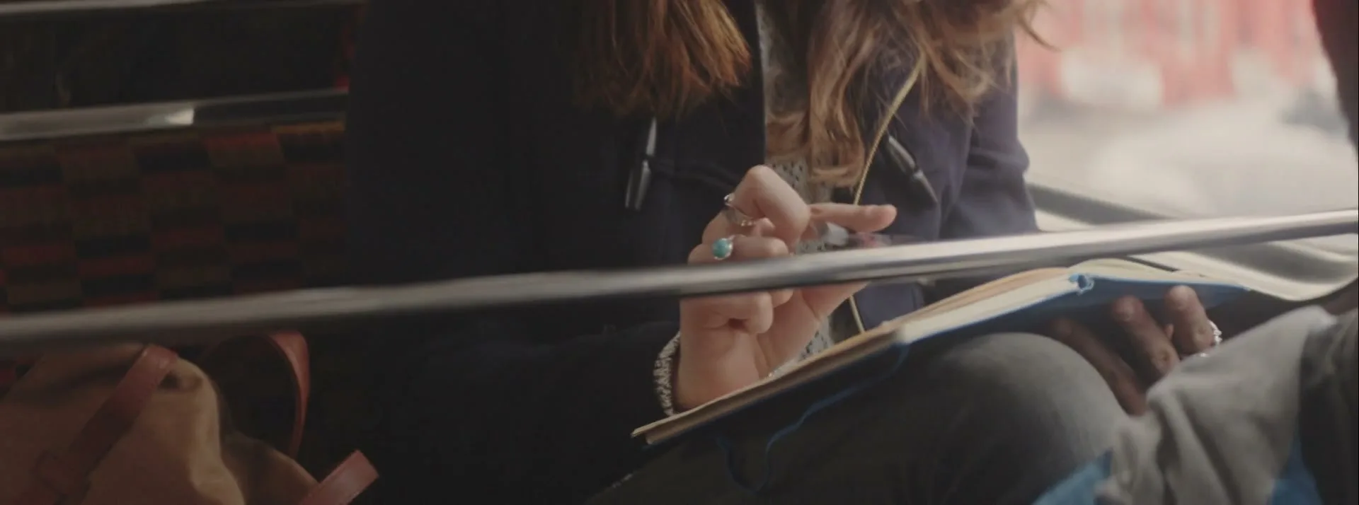 A young woman with long brown hair, wearing a dark coat and rings, holds a blue notebook and pen while riding a bus.