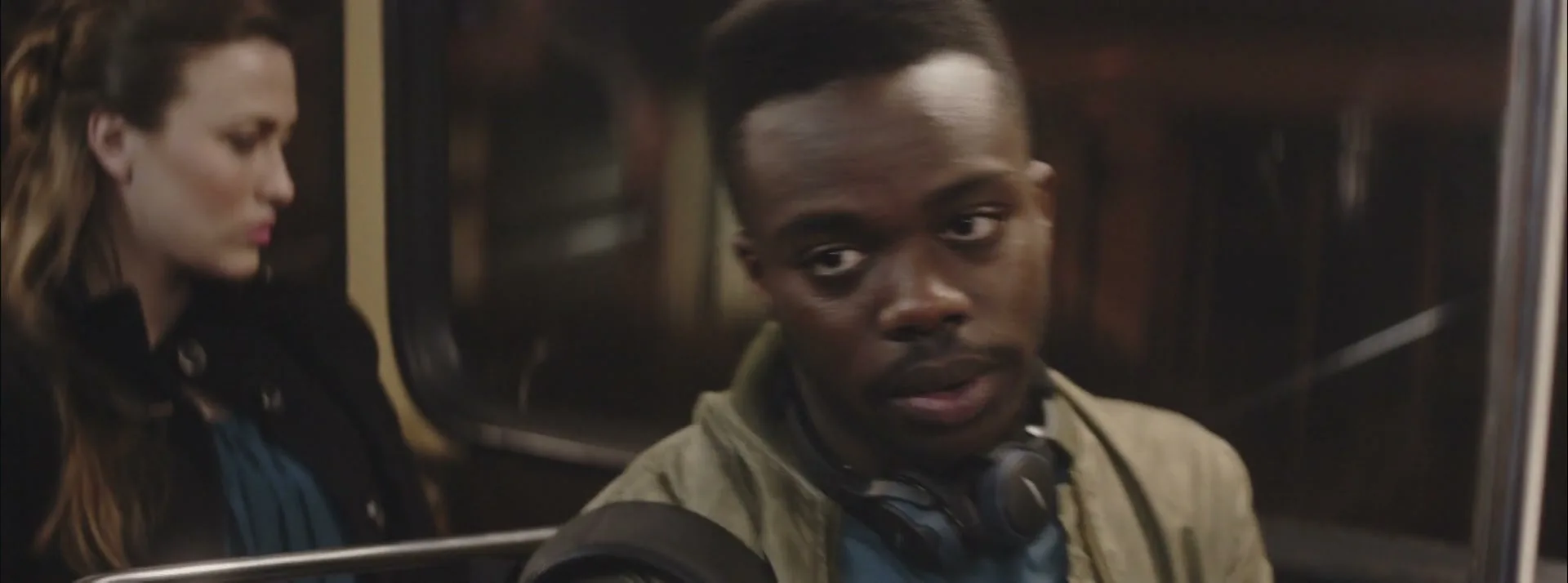 A young Black man with headphones around his neck looks around a subway car, while a young white woman sits next to him.