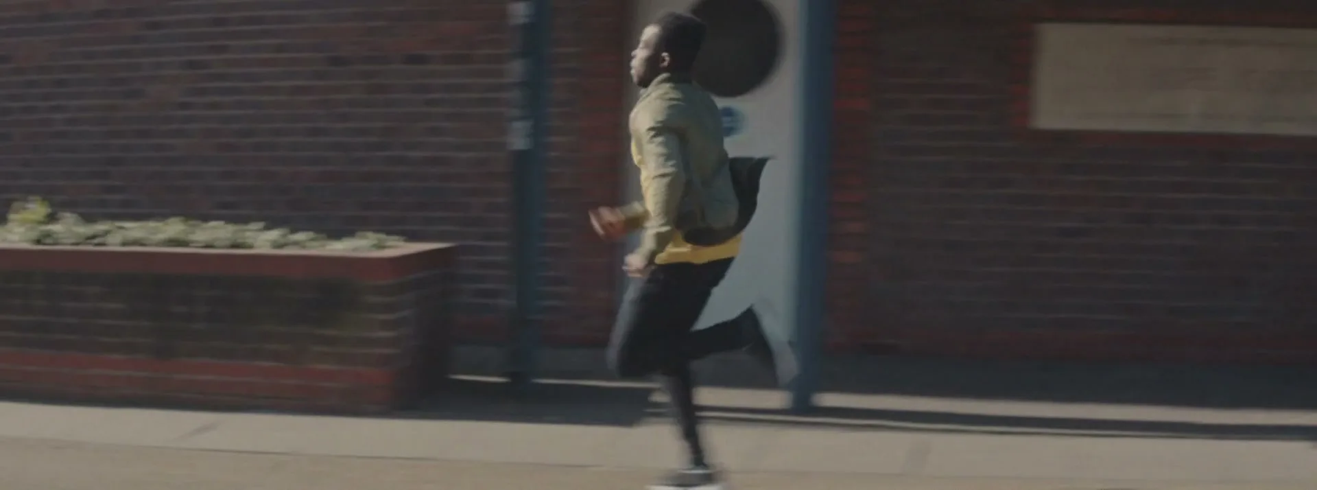 A young man in a yellow shirt and green jacket runs past a brick building on a sunny day, with a blurred car in the foreground.