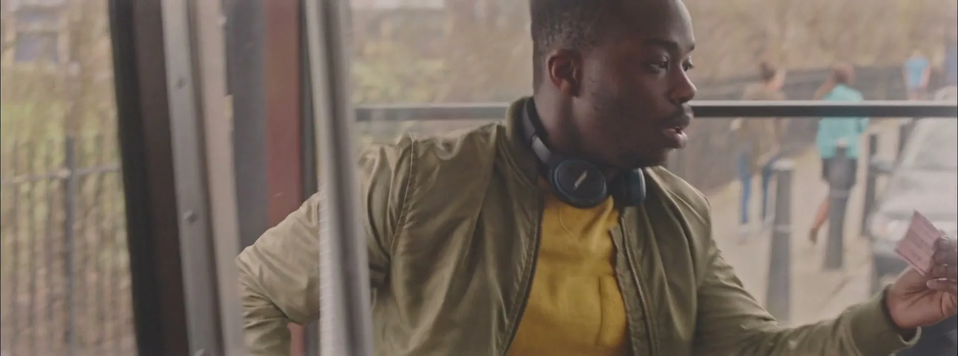 A young Black man wearing headphones around his neck looks out the window of a moving bus, holding a ticket in his hand.