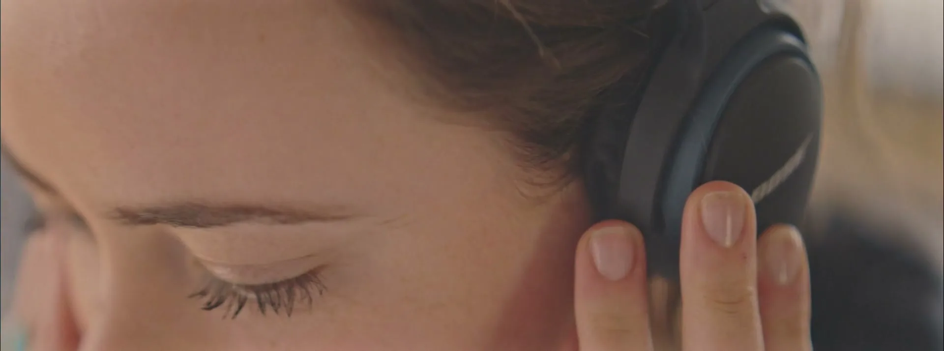 A young woman with brown hair and a Bose headphone on her ear, closes her eye while listening to music.