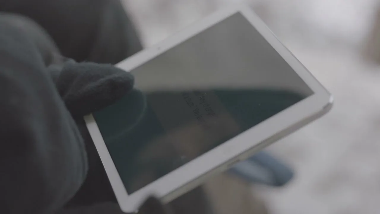 A person wearing black gloves holds a white tablet, with text appearing on screen, against a blurry, light-colored background.