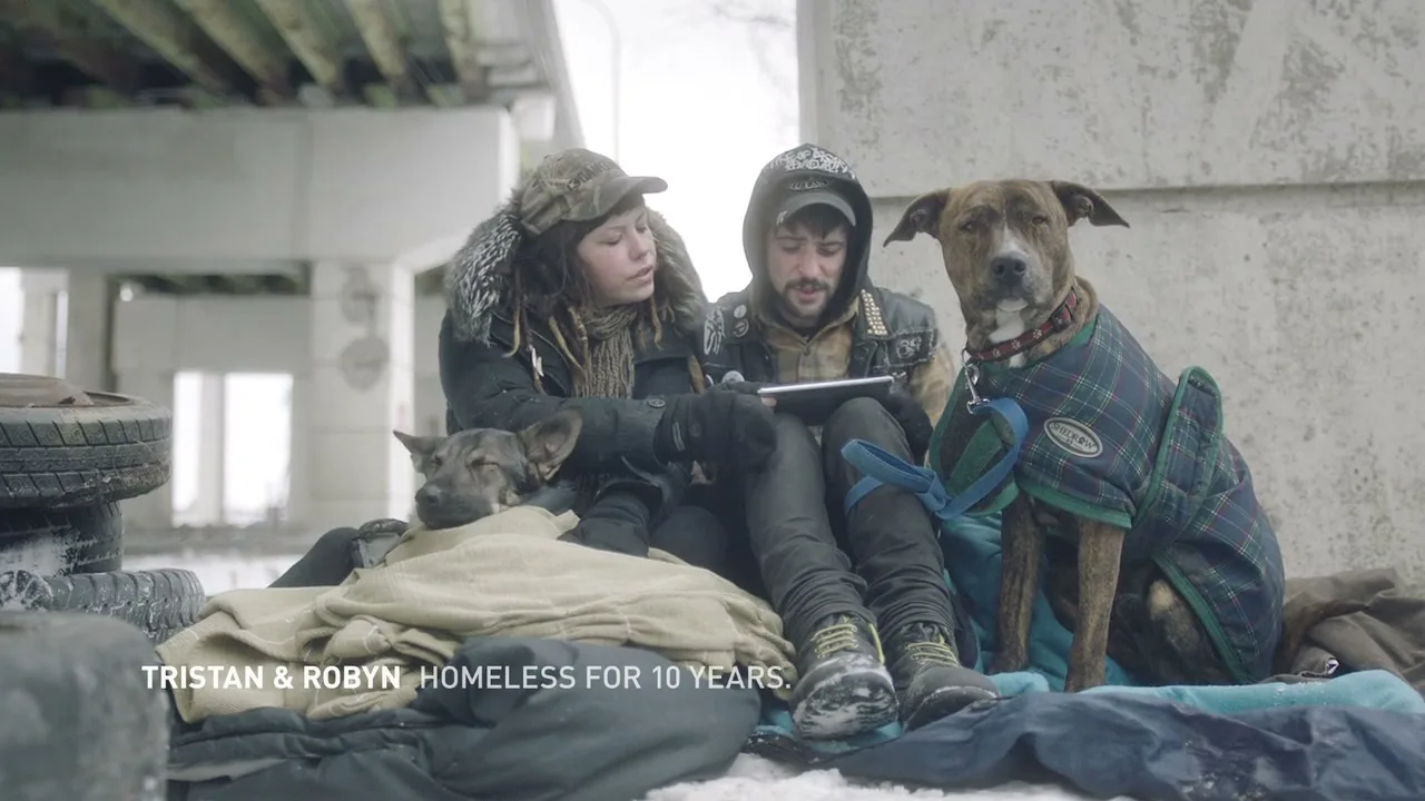 A young man and woman, dressed in winter clothes, sit on the ground with two dogs, looking at a tablet under a bridge.