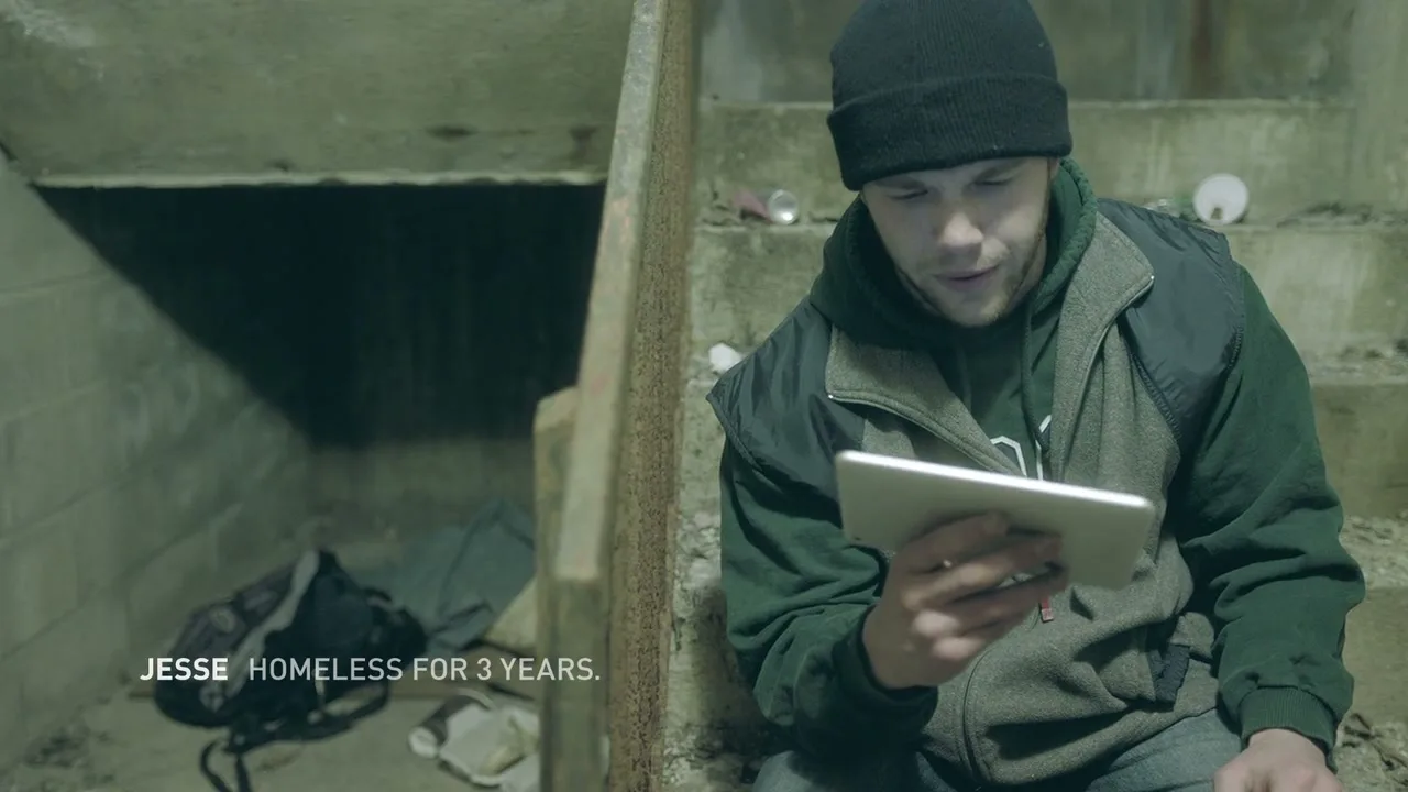 A young man, Jesse, wearing a black beanie and green hoodie, sits on concrete stairs, reading a mean tweet on a tablet in a dimly lit underpass.