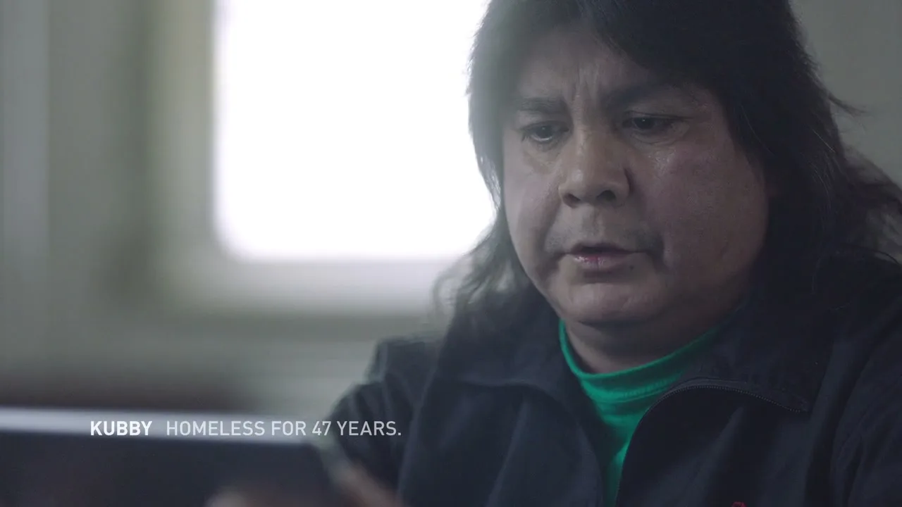 A middle-aged Indigenous man with long dark hair and a green shirt looks down sadly as he reads a mean tweet in an interior setting.