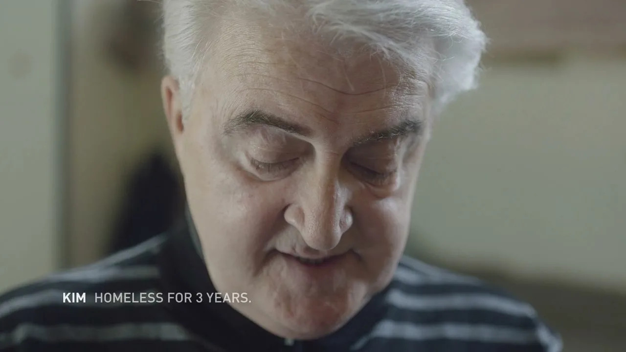 A senior man with white hair and a striped shirt reads a mean tweet about homeless people, looking down then up at the camera, indoors.
