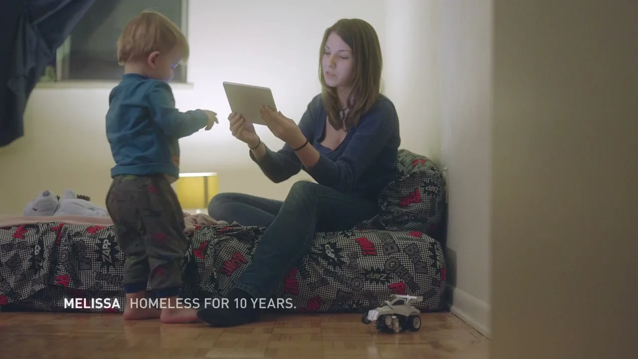 A young woman sits on a bed, looking at a tablet, while a toddler plays nearby in a dimly lit room.