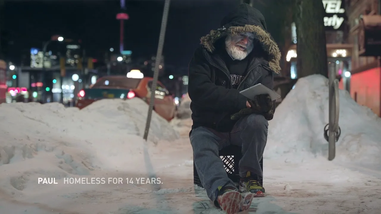 A homeless man, Paul, with a white beard and a fur-lined hood, sits on a milk crate in a snowy city street at night, holding a tablet.