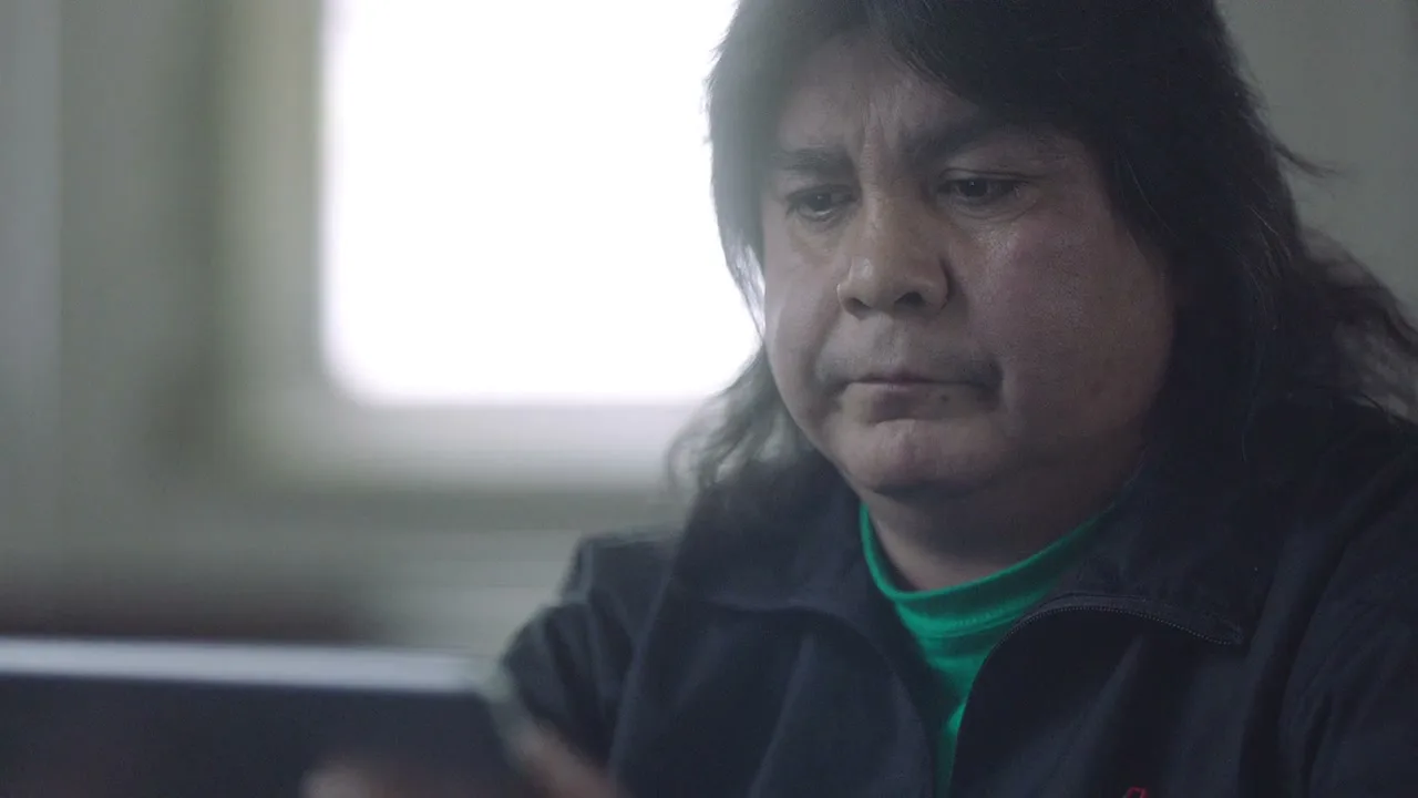 A middle-aged man with long dark hair and a green shirt looks down at a tablet with a neutral expression in an interior setting.