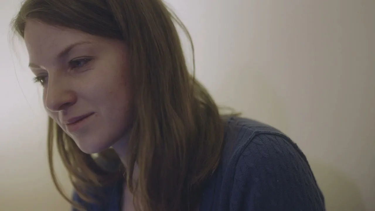 A young woman with long brown hair, wearing a blue sweater, looks down with a slight smile in an interior setting.