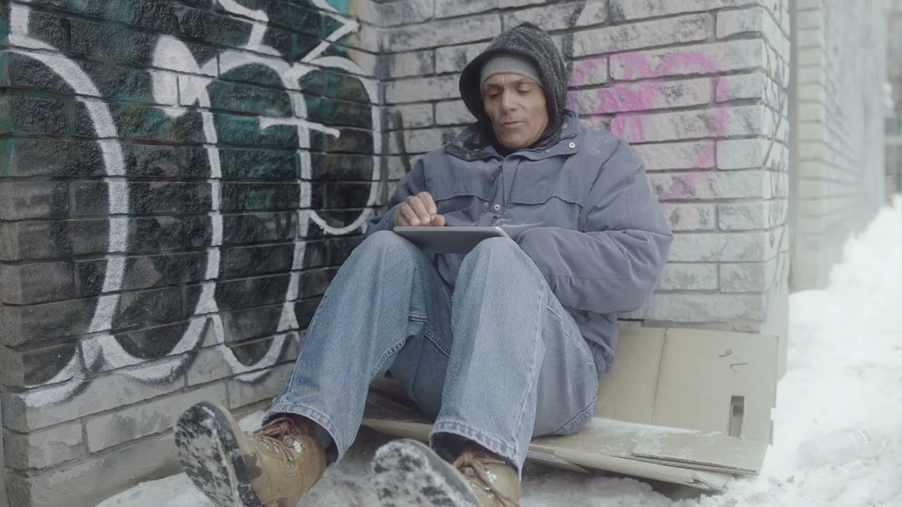 A man in his 40s, dressed in a blue jacket and jeans, sits on cardboard against a graffiti-covered wall in the snow, looking at a tablet.