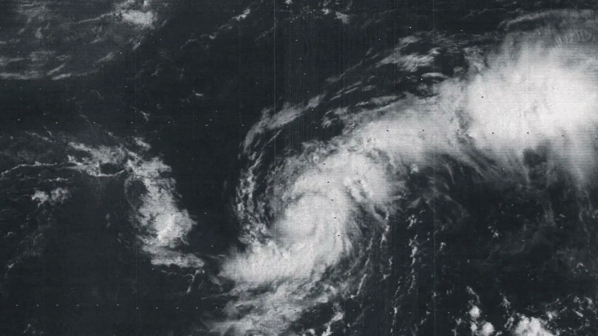 A black and white satellite image shows a swirling hurricane over the ocean, with visible static and film grain.