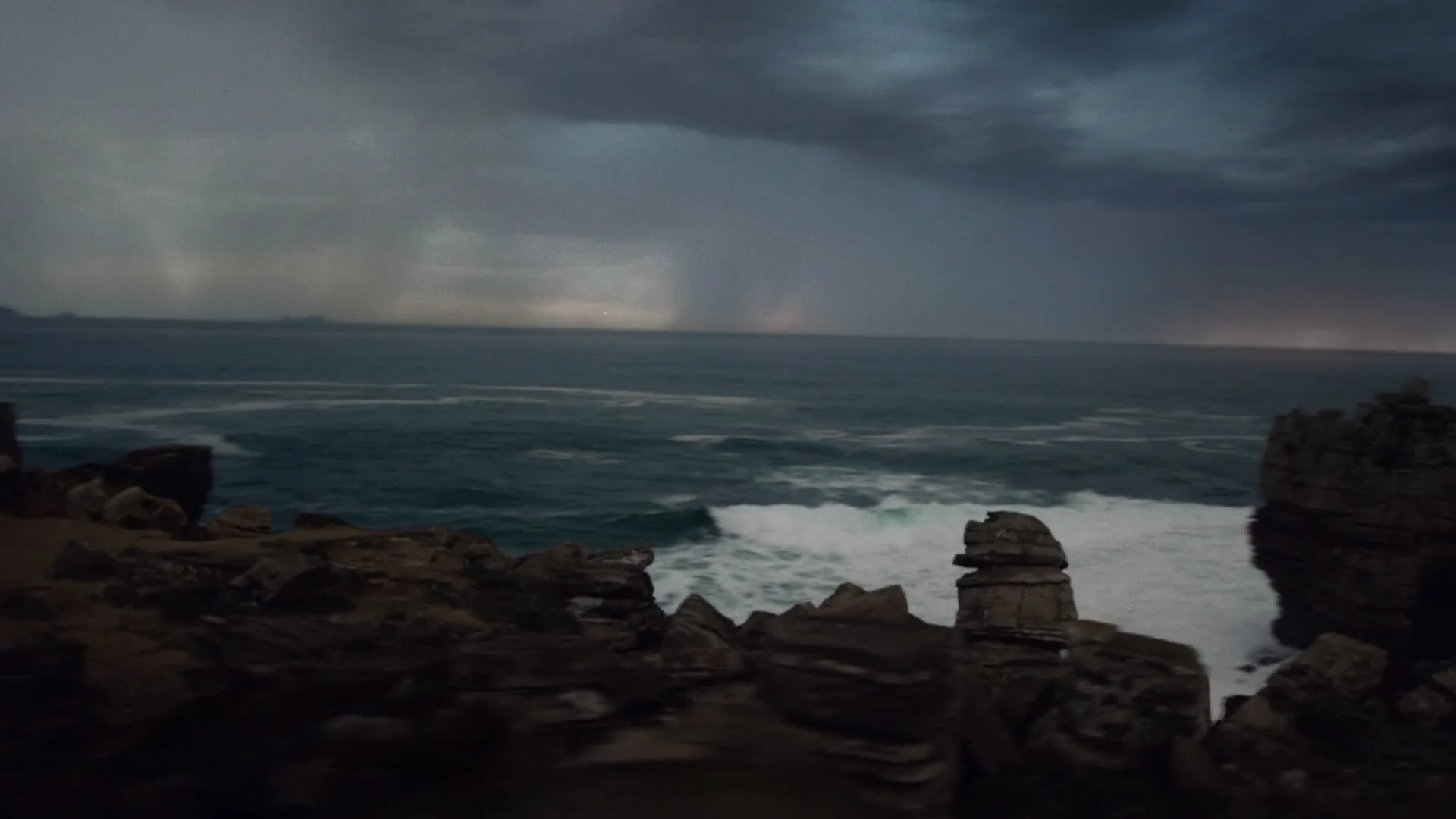 A wide shot of a stormy ocean with dark clouds and rain falling in the distance, seen from a rocky coastline.