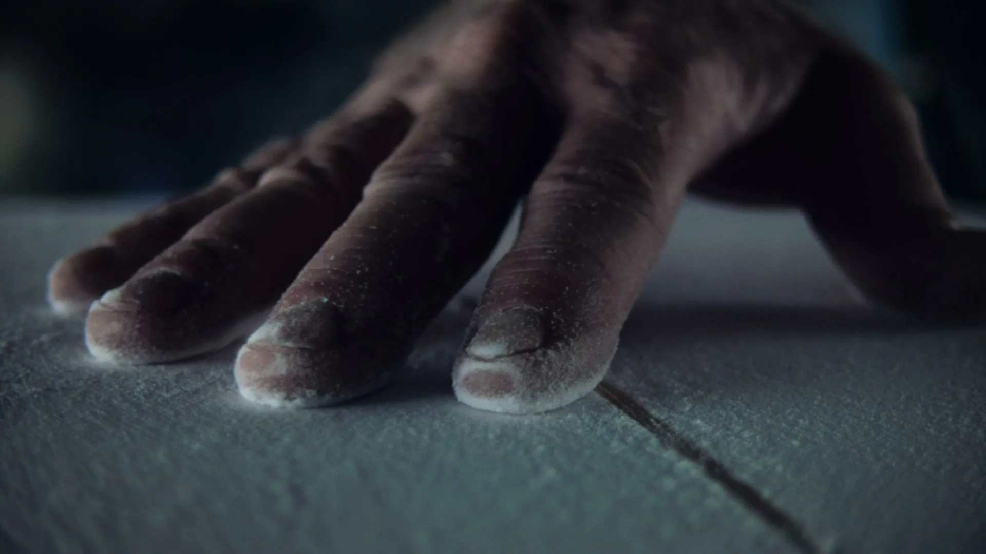A person's hand, covered in white wax, is shown in a closeup as it presses down on a surfboard in a dimly lit interior.