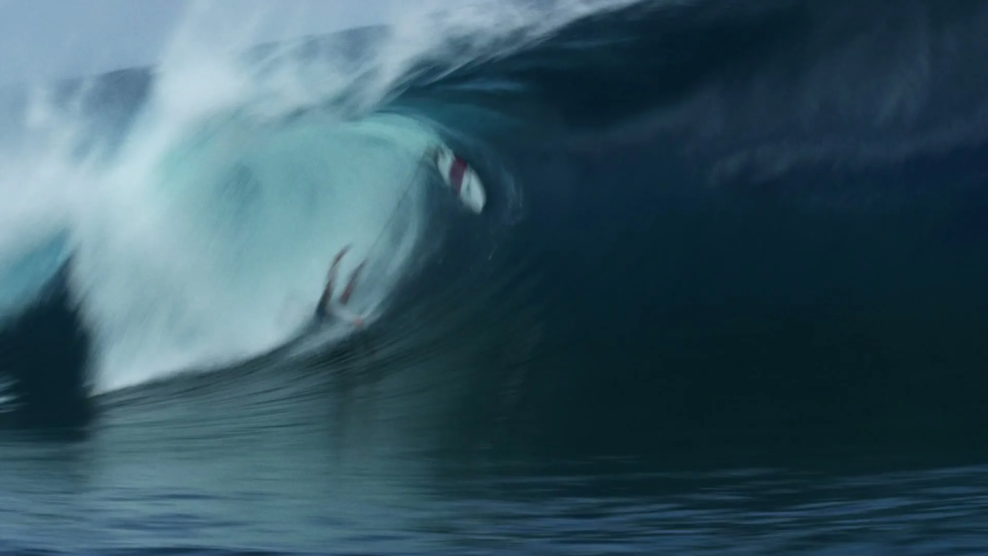 A surfer in a dark wetsuit is wiped out by a massive, breaking blue wave in the ocean under a cloudy sky.