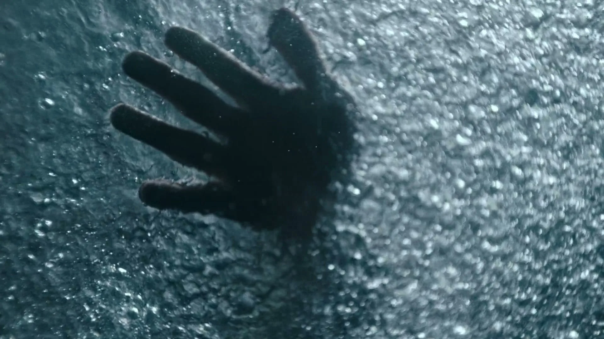A dark hand is seen through a turbulent surface of water, with many bubbles, as if submerged or pressing against a glass.